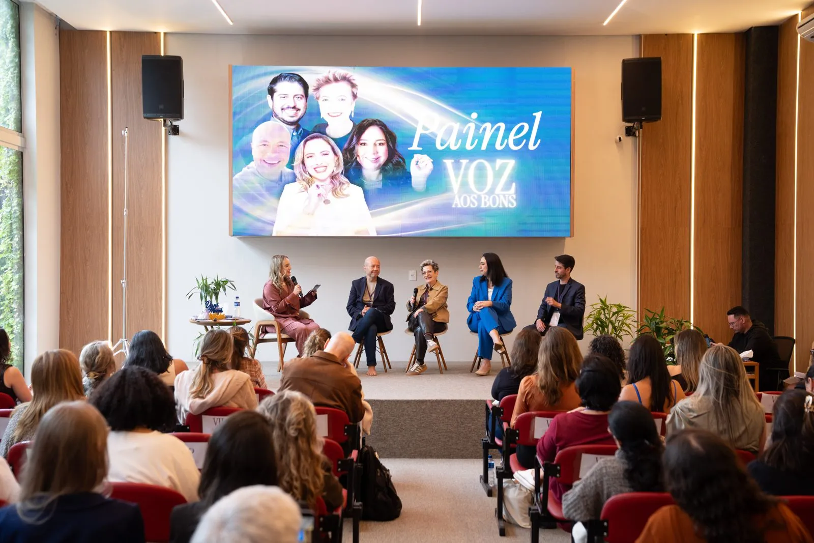 Painel com cinco convidados no palco diante de uma plateia cheia, com o telão do painel Voz aos Bons ao fundo